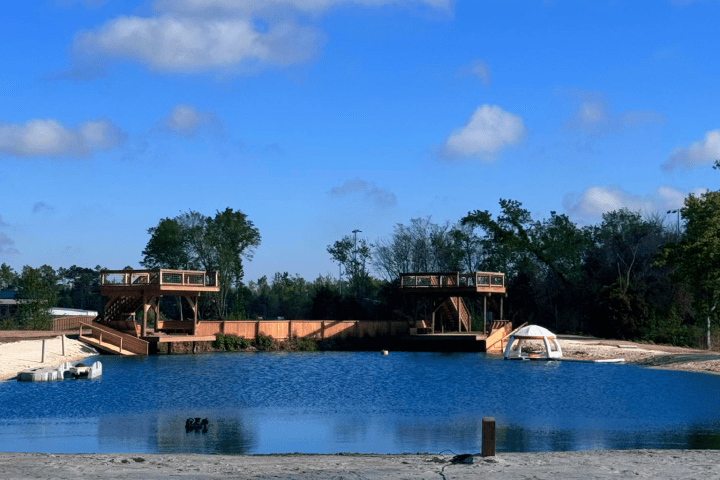Calm lake with blue sky, wooden platforms, trees, and sand in the foreground.