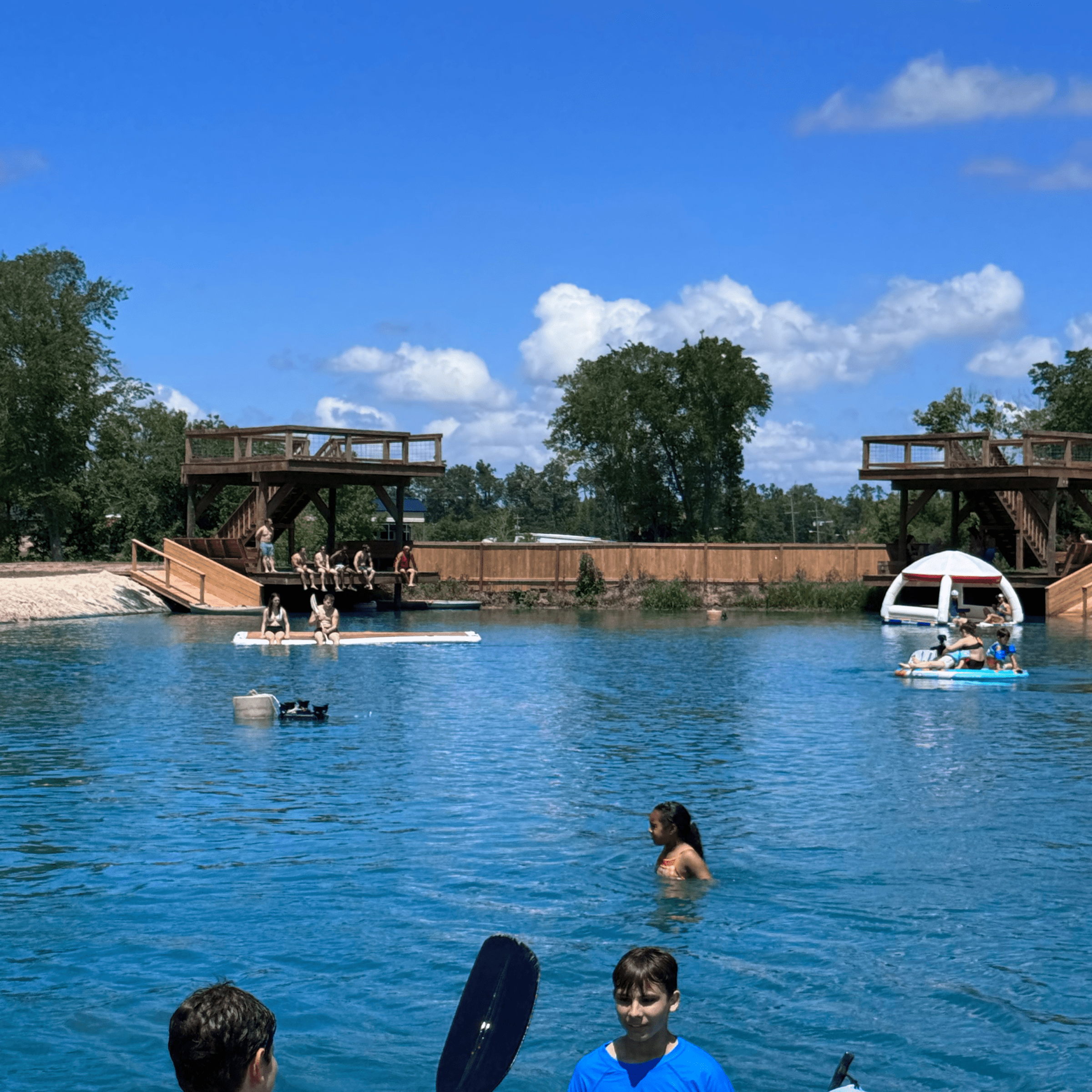 People swimming and kayaking on a lake with wooden platforms and trees in the background.