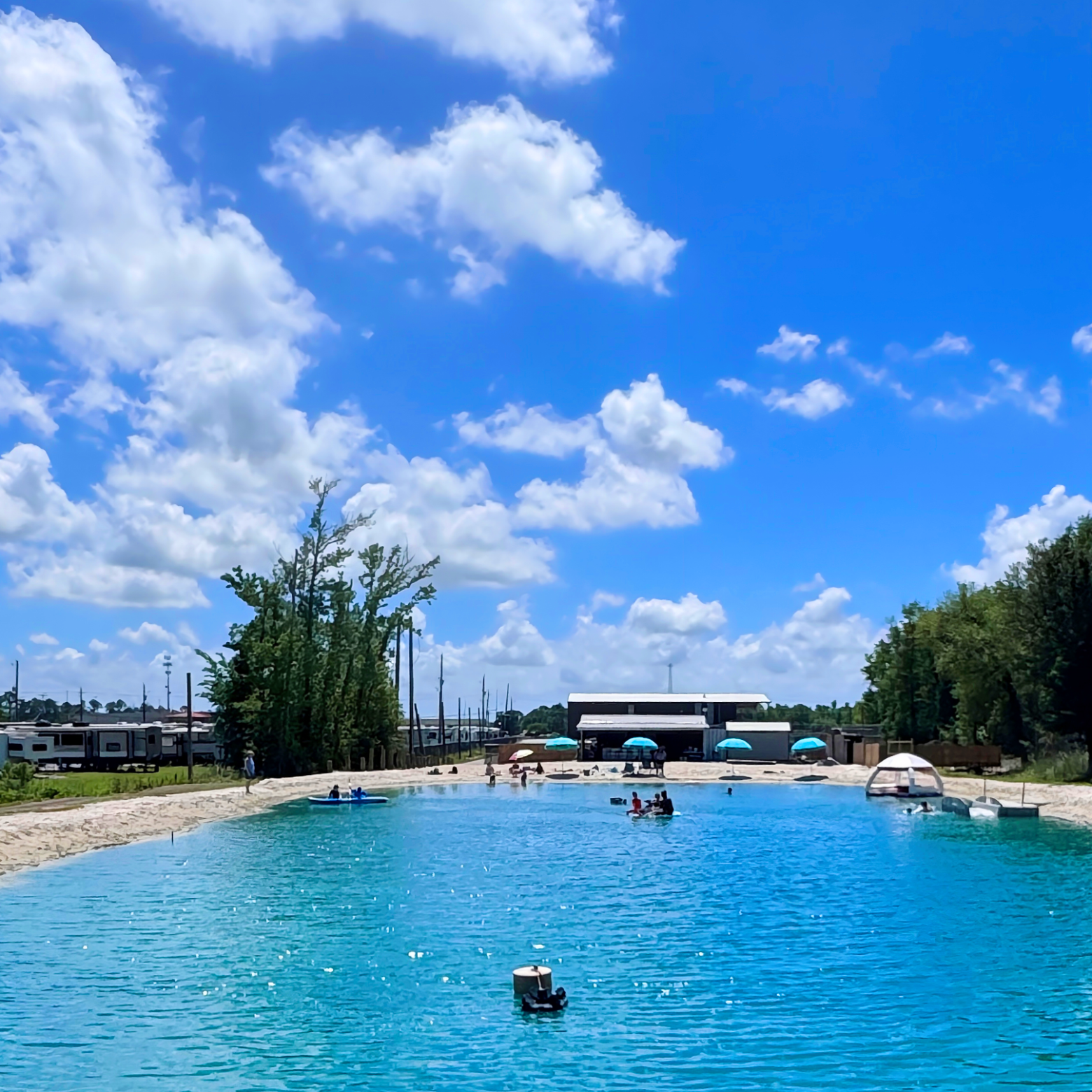People enjoying a sunny day at a large outdoor pool with inflatable rafts and surrounded by trees.