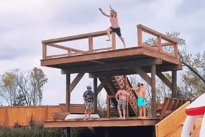 Person jumps off wooden platform into a pool, watched by three others on lower level.