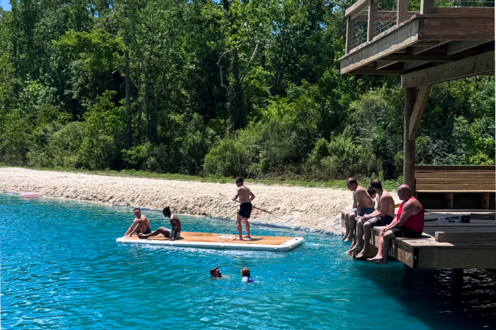 People swimming and relaxing on a dock by a clear blue lake with trees in the background.