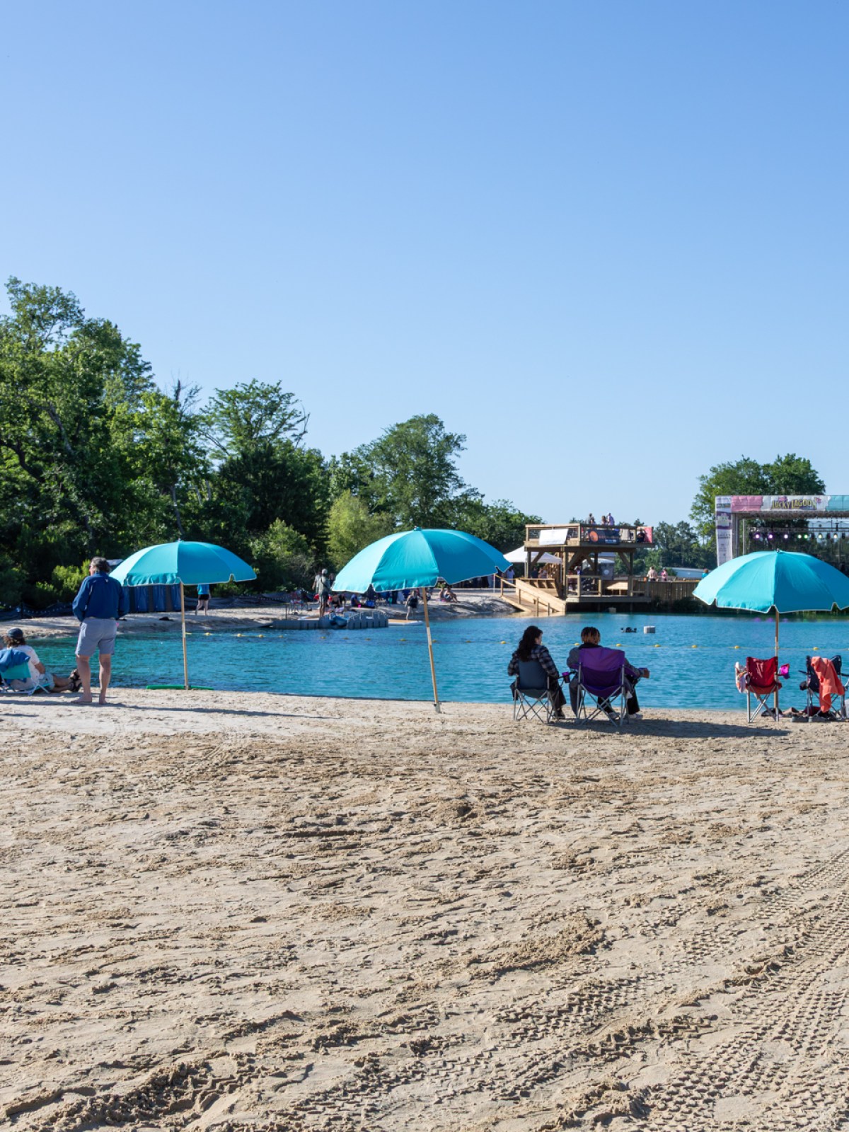 People relax on a sandy beach with blue umbrellas near a lake on a clear day.