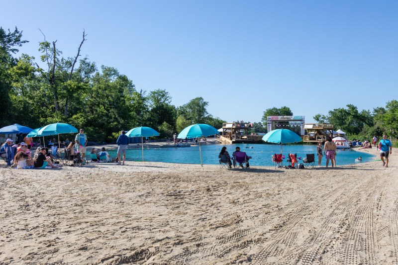 People relax on a sandy beach with blue umbrellas near a lake on a clear day.