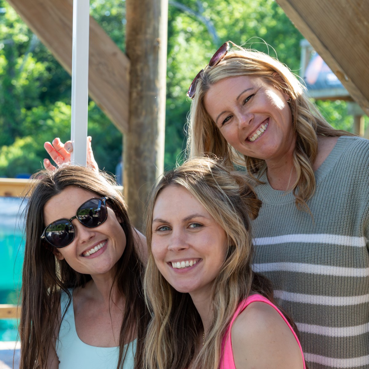 Three women smiling outdoors near a pool with greenery in the background.