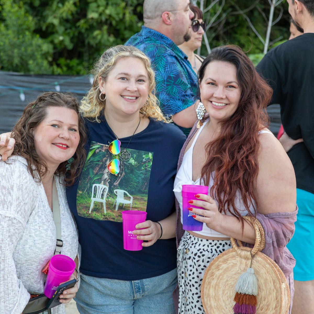 Three women smiling with drinks at an outdoor gathering.